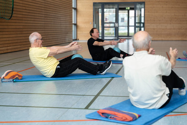 Gruppe bei einer Rückenübung im Sitzen auf Matten mit gestreckten Armen zur Kräftigung der Bauch- und Rückenmuskulatur.
