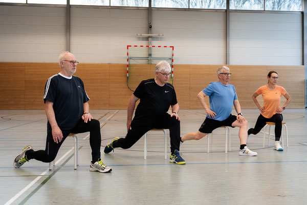 Teilnehmer des Herz- und Rehasports beim BSV Achim führen Dehn- und Kräftigungsübungen im Ausfallschritt mit Unterstützung von Hockern in der Sporthalle durch.
