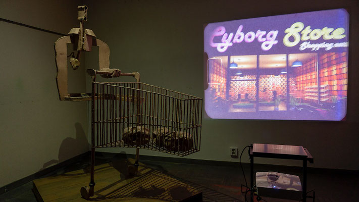 Vintage cyborg winkelkar (1960-model) — testopstelling van de sculptuur met videoprojectie van de 3D-animatie op de muur, waarbij de fysieke winkelkar en de digitale cyborg store samen één installatie vormen.