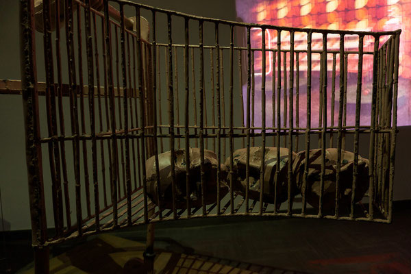 Vintage Cyborg Shopping Cart (1960 model) — detail view of a metal shopping cart containing multiple sculptural heads lying inside the basket, shown within a dark exhibition setup with projected light in the background.