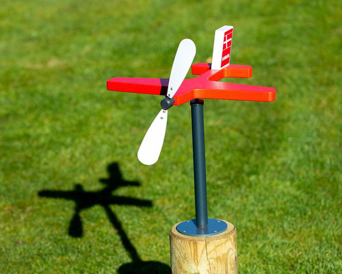 Ein Windspiel in Flugzeugform in den dänischen Nationalfarben rot und weiß. Das Windspiel ist auf einem runden Holzpfosten befestigt und steht vor einer grünen Rasenfläche.
