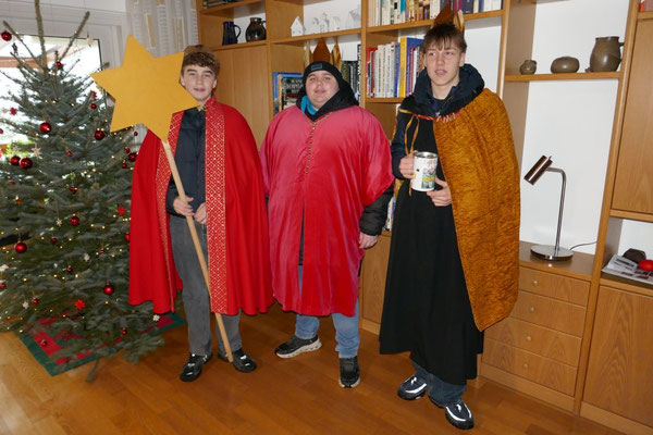 Drei Sternsinger stehen vor dem Weihnachtabaum und einer Bücherwand, um ihr Sternsingerlied zu singen und den Segen zu übergringen.