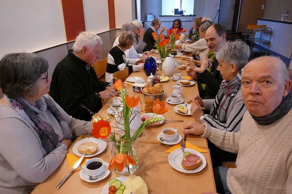 Foto zeigt Blick während des gemeinsamen Frühstücks in den Gemeindesaal. Die Teilnehmenden sitzen an einer langen Tischreihe und haben das Früstück vor sich stehen.