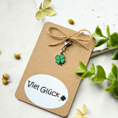 Glückwunschkarte mit Kleeblatt-Anhänger und “Viel Glück” Text auf weißem Oval, umgeben von Pflanzen.