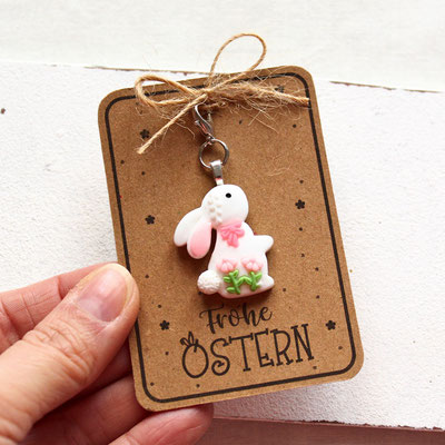 Weiße Karte mit „Frohe Ostern“-Schriftzug und Hasenanhänger, dekoriert mit rosa Blumen, Schleife und grünem Blattwerk.