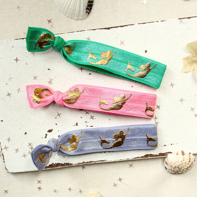 Drei Haargummis mit goldenen Meerjungfrauenmotiven auf grünem, rosa und lila Band, dekoriert mit Muscheln und Sternen.