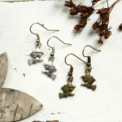 Zwei Paar Ohrringe mit Hexenmotiv, eines silbern, das andere bronzefarben, neben getrockneten Blumen und Blattfragmenten.
