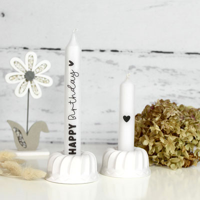 Zwei Kerzen auf Kerzenhaltern mit ‘Happy Birthday’ Text und einer getrockneten Blume.