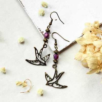 Bronzefarbene Vogel-Ohrringe mit lila Perlen auf hellem Untergrund mit getrockneten Blumen.