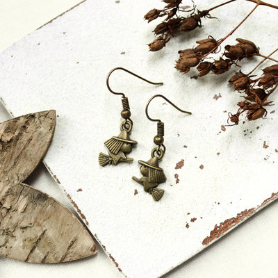 Bronze Hexen Charm Ohrringe neben getrockneten Blumen und einem Stück Holz auf hellem Untergrund.