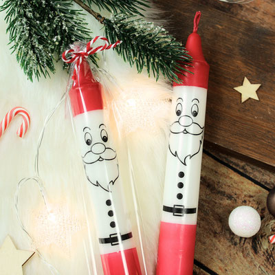 Zwei Weihnachtskerzen in Form von Weihnachtsmännern, umgeben von Tannenzweigen, Zuckerstangen und Weihnachtsdekorationen.