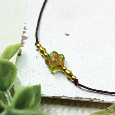 Handgefertigtes Fußband mit grün-roter Glasperle und goldenen Akzenten auf weißem Hintergrund.