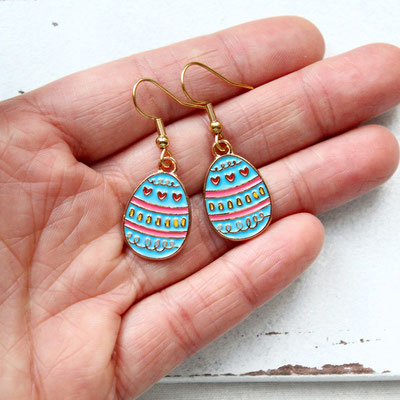 Hand hält bunte Ohrringe in Eiform mit blauen, roten und gelben Mustern und goldenen Haken.