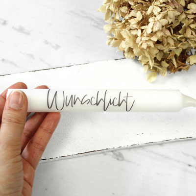 Hand hält weiße Stabkerze ‘Wunschlicht’ vor trockenen Blumen.