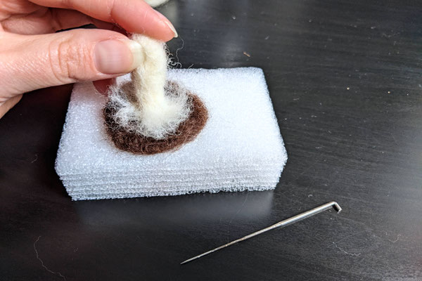 Hand putting mushroom stem onto the cap, on foam block and felting needle in the foreground