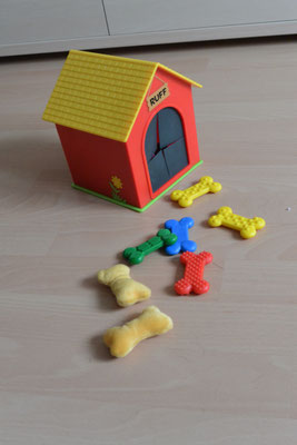 Eine sehr kleine Plastik-Hundehütte mit einem Eingriff für die Hand. Dazu gehören Plastik- und Stoffknochen mit unterschiedlichen Oberflächen, z.B. weich, glatt, knubbelig etc. Immer zwei sind gleich. 