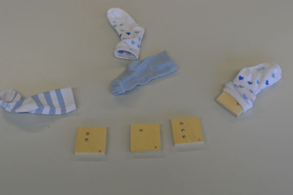 Auf dem Tisch liegen Holzklötzchen, auf denen mit Nägeln die Punktschriftbuchstaben b, a und l dargestellt sind. Daneben liegen Socken, in einem Socken sieht man einen weiteren Holzklotz stecken. 