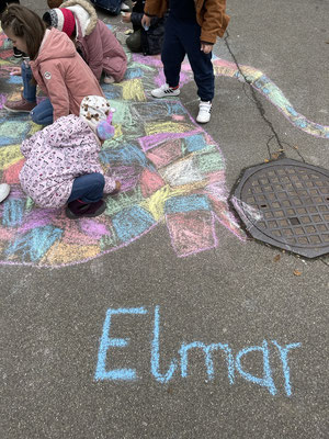 Kinder malen mit Kreide auf dem Schulhof. Es ist ein blauer Schriftzu mit "Elmar" zu lesen.