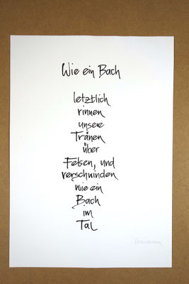 Vollansicht Kalligrafie „Wie ein Bach“ Tusche auf Papier vor Hintergrund einer Tischplatte