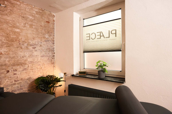 Behandlungsraum: Fensterfront mit Jalousie und PLACE ØNE-Logo, hell und freundlich; im Vordergrund die Therapieliege im PLÆCE ØNE Therapy CoWorking Hamburg