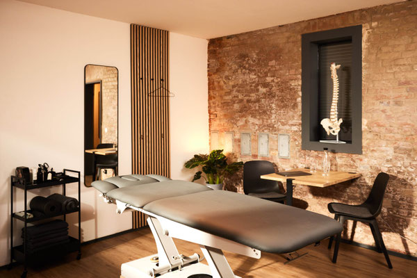 Zweiter Blick in den Behandlungsraum mit Spiegel, Tisch, Grünpflanze, kleinem Anatomiemodell und Therapieliege vor warmer Backsteinwand; ruhige, freundliche Atmosphäre im PLÆCE ØNE Therapy CoWorking Hamburg