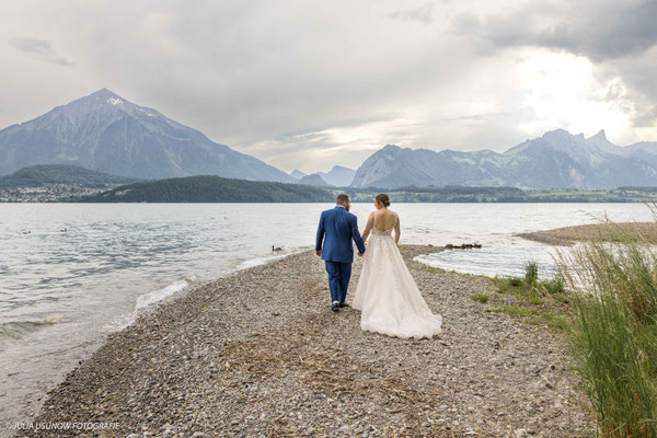 Brautpaar läuft Hand in Hand auf einer Sandbank vor dem Parkhotel Gunten im Berner Oberland, Berge im Hintergrund