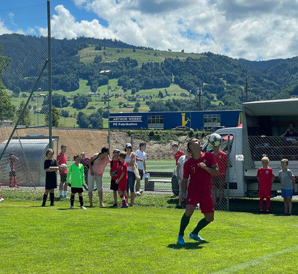 Fußball-Freestyler mit Ball am Spielfeldrand vor Zuschauern und Schweizer Hügellandschaft