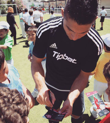 Mann in schwarzem Adidas-Shirt unterschreibt Autogrammkarten, während Kinder um ihn herum Karten hinhalten.