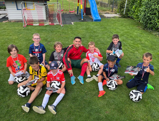 Children sitting in a garden after a mini tournament, holding medals and footballs