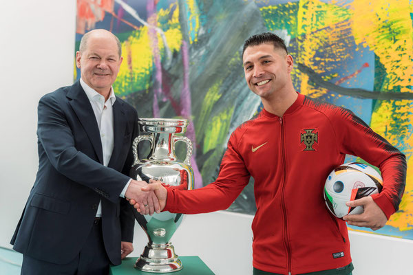 Saki im roten Fußballoutfit beim Handschlag neben dem EM-Pokal in einem Innenraum des Bundeskanzleramts