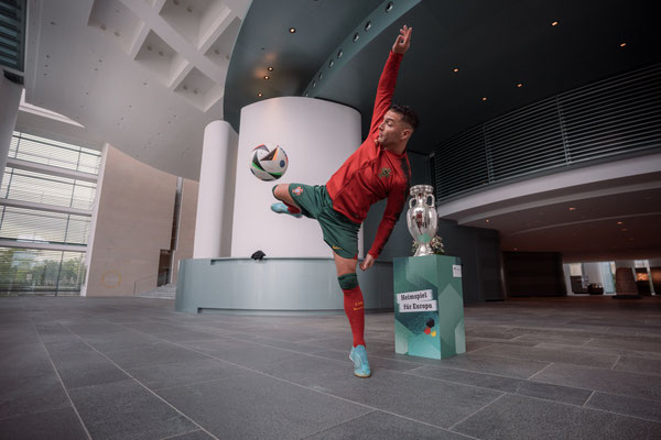 Fußball-Freestyler Saki zeigt eine Freestyle-Pose mit Ball im großen Innenraum des Bundeskanzleramts neben dem EM-Pokal