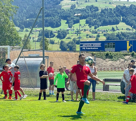 Fußball-Freestyler balanciert Ball auf dem Bein vor Kindern und Alpenkulisse in der Schweiz