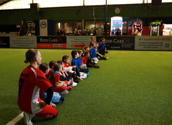 Kinder sitzen auf Kunstrasen in der Indoor-Soccerworld Berlin und warten auf das Fußballtraining