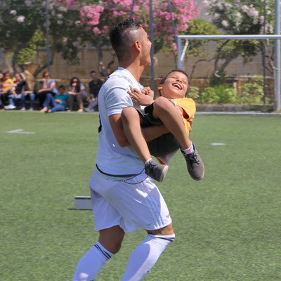 Fußball-Freestyler Saki trägt ein lachendes Kind auf dem Kunstrasenplatz der St Clare College Primary School Malta.