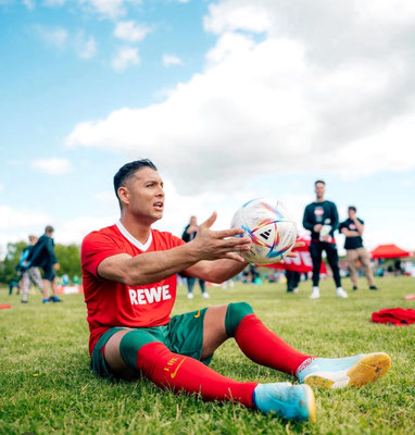 Saki sitzt auf einer Wiese und hält einen Fußball bei einem REWE-Outdoor-Event.