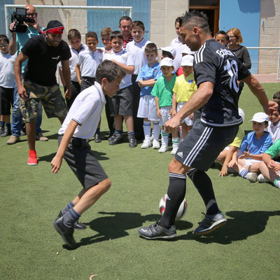 Mann in schwarzer Sportkleidung dribbelt mit dem Fußball im 1-gegen-1 gegen einen Schüler; Kinder schauen zu.