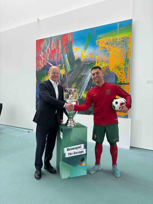 Saki und Bundeskanzler Olaf Scholz im Anzug stehen neben dem EM-Pokal auf Podest im Bundeskanzleramt in Berlin