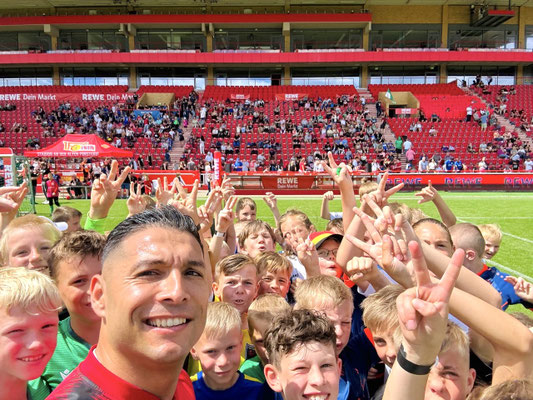 Selfie von Saki mit vielen Kindern vor roten Stadiontribünen bei einem REWE-Event