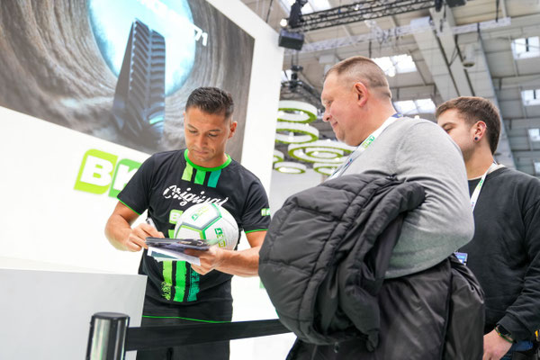 Saki gibt einem Besucher am BKT-Stand in Hannover ein Autogramm und hält dabei einen BKT-Fußball