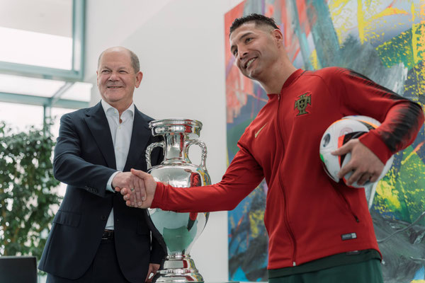 Nahaufnahme von Saki beim Handschlag neben dem EM-Pokal in einem hellen Raum des Bundeskanzleramts