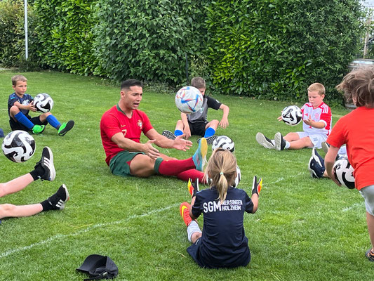 Fußball-Workshop mit Saki und Kindern bei einer Sitzübung mit Bällen auf dem Rasen in Bayern