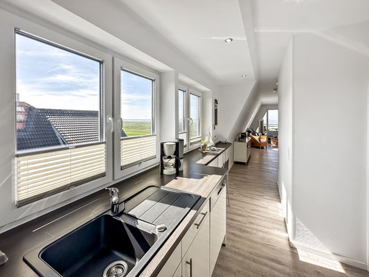 Eine moderne Küche mit weißen Schränken, einer großen Arbeitsplatte, einer Spüle und Küchengeräten. Große Fenster sorgen für natürliches Licht und bieten eine atemberaubende Küchenzeile mit Meerblick. Der Raum geht nahtlos in den Wohn- u. Essbereich über.