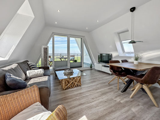Das Penthouse Dünenweg 37 verfügt über ein helles, modernes Wohnzimmer mit schrägen Decken. Der Wohn- und Essbereich besteht aus einem Sofa, einen Fernseher, einen Essbereich und großen Fenstern und dem Zugang zur Loggia mit atemberaubender Meerblick.