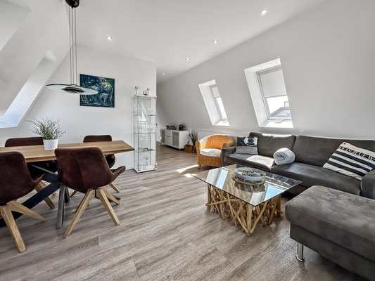  Modernes Wohnzimmer in der Ferienwohnung Cuxhaven mit Dachschräge, einem Esstisch mit vier Stühlen, einem Ecksofa und einem Sessel. Durch die zahlreichen Fenster hell und verfügt aus fast allen Wohnbereichen über Meerblick.