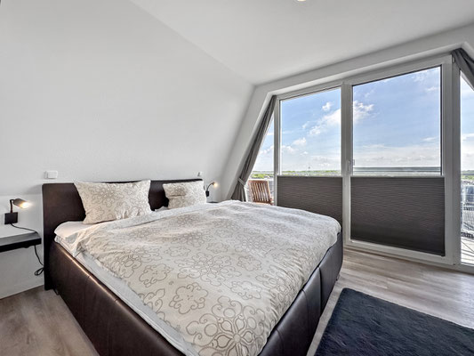 Das Schlafzimmer mit einem großen Boxspringbett. Das Fenster über die gesamte Wand bietet einen atemberaubenden Blick nach draußen und bietet den Zugang zum Balkon.