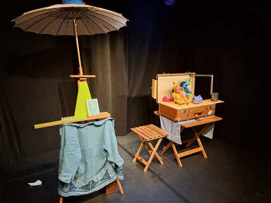 Das Köfferchen steht auf einem Holztisch. Daneben ein Holzhocker und ein Ständer mit einem aufgespannten Regenschirm.