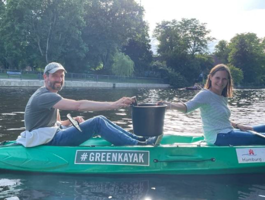 Olaf und Sandra sitzen im Green Kayak und zeigen ihren Eimer mit gesammeltem Müll