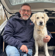 Thomas sitzt im geöffneten Kofferraum eines Fahrzeugs. Er trägt eine dunkelblaue Jacke und eine blaue Jeans. Neben ihm sitzt ein mittelgroßer, hellbeiger Hund mit lockigem Fell. Thomas hält den Hund mit einer Hand am Halsband fest.