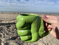 Eine Hand hält eine grüne Tasse ins Bild. Die Tasse ist eine dreidimensionale Hulk-Faust. Im Hintergrund ist ein Strand und Meer zu sehen.