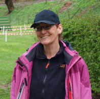Sybille trägt schwarze Baseballkappe, schwarzen Fleecepullover mit orangem Reißverschluss und pinke Outdoorjacke, steht auf einer Wiese mit Zaun im Hintergrund.